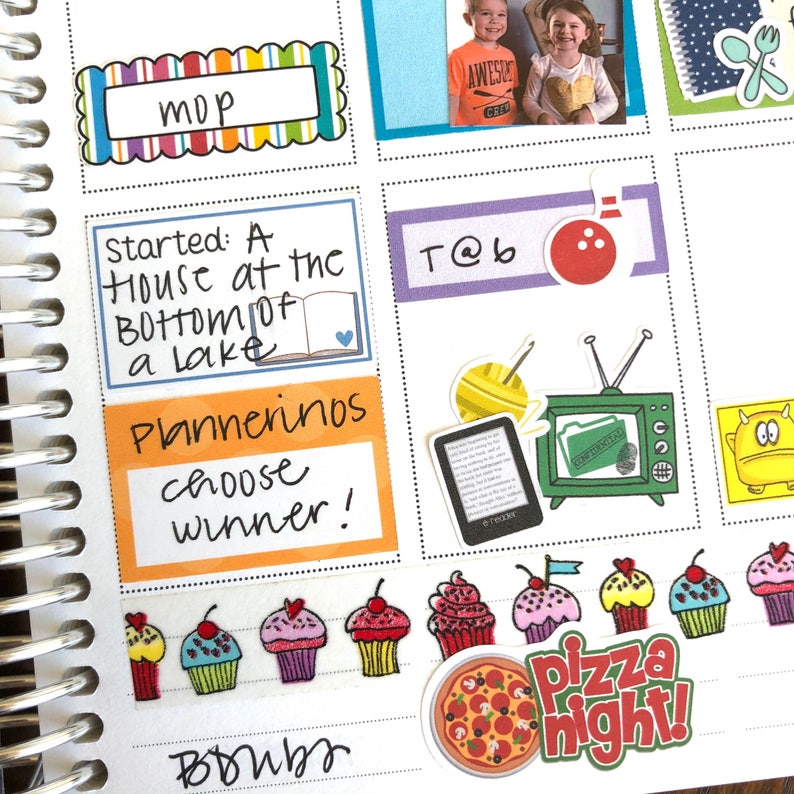 May include: A planner page with colorful stickers and handwritten notes. The stickers include a book, a bowling pin, a television, a cupcake, and a pizza slice. The handwritten notes include "Started: A house at the bottom of a lake", "Plannerinos", "choose winner!" and "T@b".
