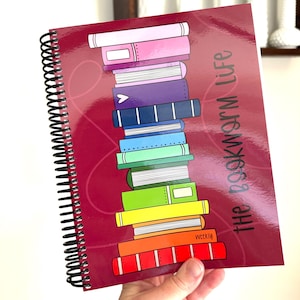 May include: A spiral-bound notebook with a dark red cover featuring a vertical stack of colorful illustrated books. The text "the bookworm life" is printed in dark gray on the right side. The books are in shades of pink, purple, blue, green, yellow, orange, and red.