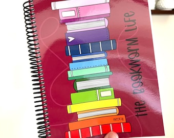 May include: A spiral-bound notebook with a dark red cover featuring a vertical stack of colorful illustrated books. The text "the bookworm life" is printed in dark gray on the right side. The books are in shades of pink, purple, blue, green, yellow, orange, and red.