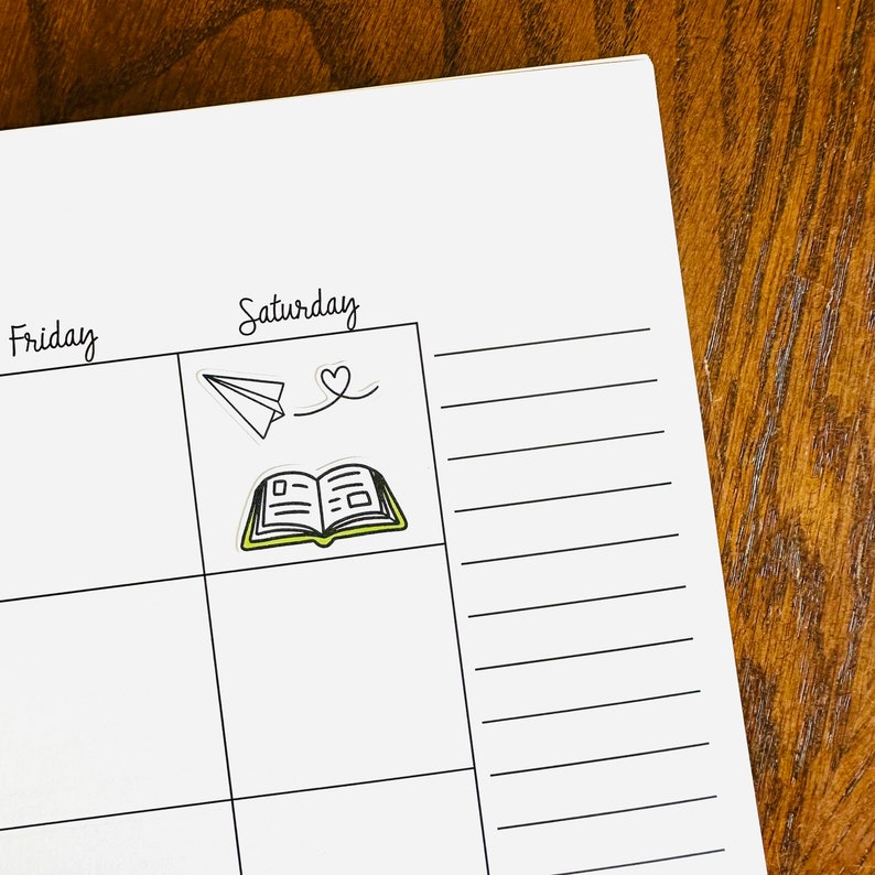 May include: A white planner with black lines and the words "Friday" and "Saturday" written on it. There are two stickers on the planner, one of a paper airplane and one of an open book.