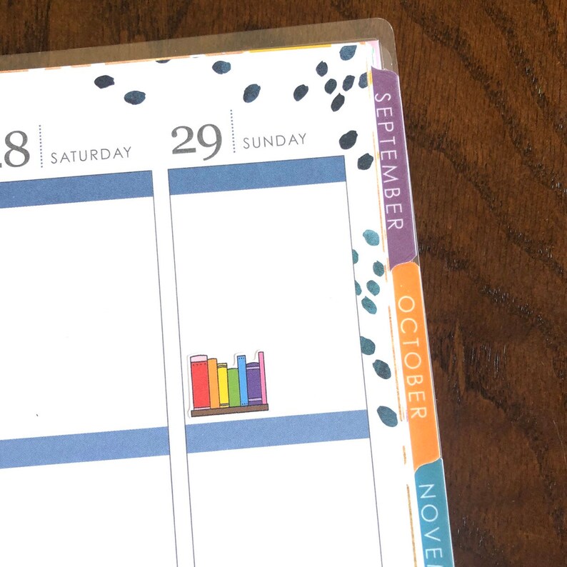 May include: A close-up of a planner with colorful tabs for September, October, and November. The planner has a black and white polka dot pattern and a sticker of a bookshelf with rainbow colored books.