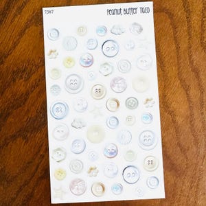 May include: A sheet of stickers featuring various sizes and styles of buttons in shades of white, cream, and pastel colors. Some buttons have iridescent finishes. The text "PEANUT BUTTER TACO" is printed at the top.