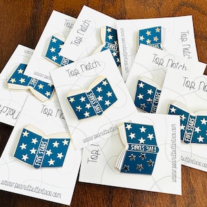 May include: Enamel pins in the shape of an open book with a blue cover and gold trim. The book's pages are filled with white stars, and the text "FIVE STARS" is displayed. Each pin is on a white card with "Top Notch" printed on it.