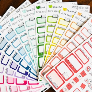 May include: A collection of colorful sticker sheets featuring various rectangular labels with rounded corners. Each sheet includes small book icons, the text "book club," "bookish," and "bookish event," arranged in a fan pattern with shades of red, orange, yellow, green, blue, and purple.