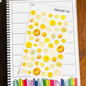 May include: A sheet of yellow and cream-colored buttons in various sizes and shapes, including stars, butterflies, and swirls. The sheet is labeled "Peanut Butter Taco" and "7382". The buttons are arranged on a lined notebook page.