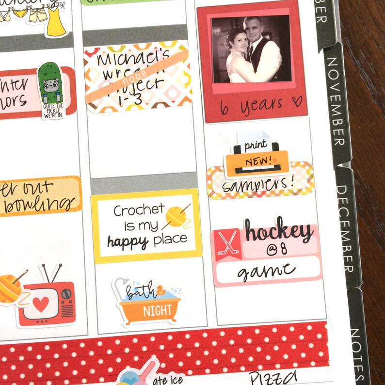 Peut inclure: Une page d'agenda avec des autocollants color&eacute;s et des notes manuscrites. Les autocollants comprennent une photo d'un couple, un crochet et de la laine, une baignoire, une crosse de hockey et un palet, et une imprimante. Les notes manuscrites comprennent "6 ans", "imprimer nouveau ! &eacute;chantillons !" et "hockey @8 jeu".