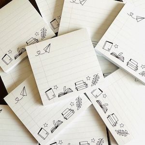 Könnte beinhalten: Ein Stapel weißes, liniertes Notizblockpapier mit schwarzen Tintenillustrationen von Büchern, Sternen, Blättern und Papierflugzeugen. Das Notizblockpapier hat ein liniertes Design.