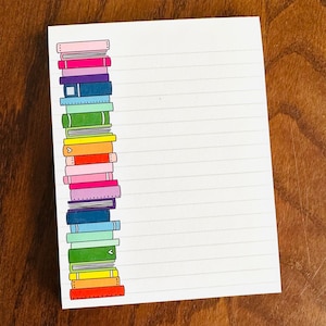 May include: A white notepad with horizontal lines and a colorful illustration of a stack of books on the left side. The books are in various colors, including pink, purple, blue, green, orange, and yellow.