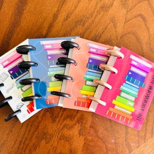 May include: Four small disc-bound notebooks with colorful illustrations of stacked books on the covers. The notebooks feature black and metallic discs along the spines. The rightmost notebook cover includes the text "the bookworm life".