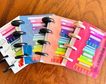May include: Four small disc-bound notebooks with colorful illustrations of stacked books on the covers. The notebooks feature black and metallic discs along the spines. The rightmost notebook cover includes the text "the bookworm life".