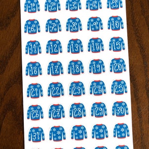 May include: Thirty-one blue and white Christmas sweater stickers with snowflake designs and white numbers 1 through 31. The stickers are on a white sheet of paper with the words "Peanut Butter Taco" written in black at the top.