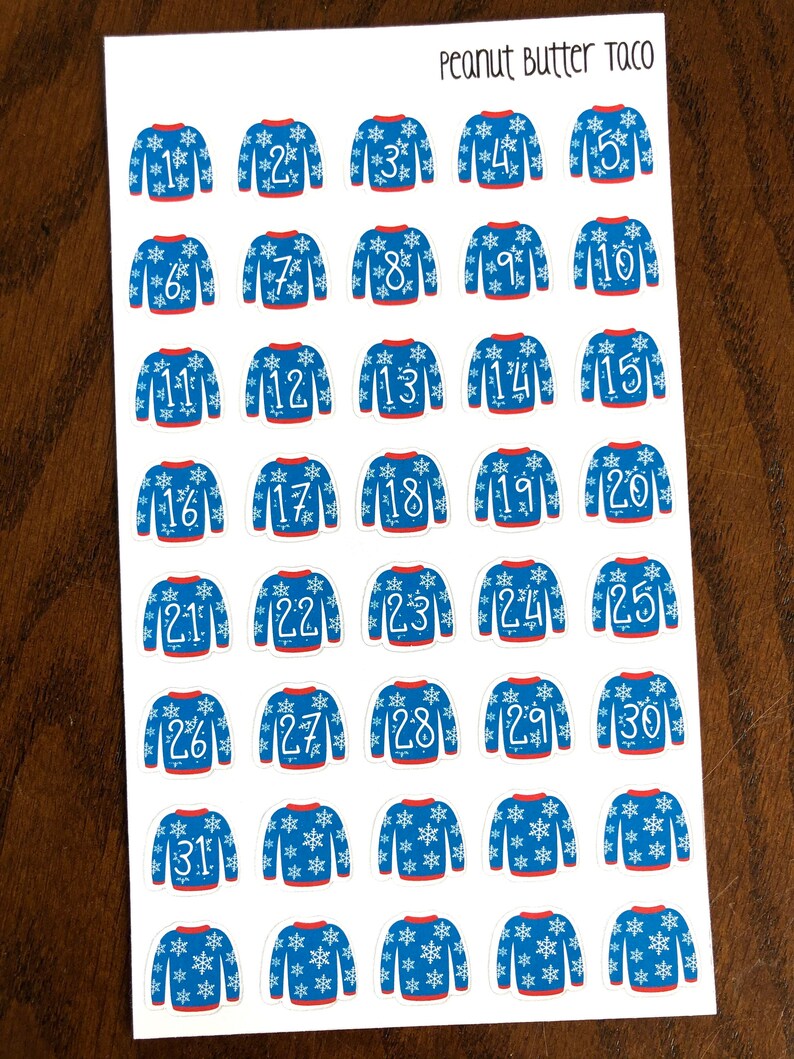 May include: Thirty-one blue and white Christmas sweater stickers with snowflake designs and white numbers 1 through 31. The stickers are on a white sheet of paper with the words "Peanut Butter Taco" written in black at the top.