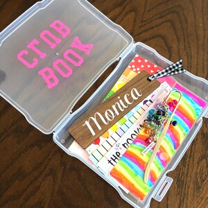 May include: A clear plastic storage box with the words "Scrap Book" written in pink on the lid. The box is open and contains a variety of colorful papers, a wooden bookmark with the name "Monica" written on it, and a gold bookmark with colorful beads.