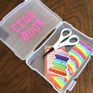 May include: A clear plastic storage box with the words "Story Book" written in pink letters on the lid. Inside the box are a pair of scissors, a rainbow-colored bookmark, and a red card with a stack of colorful books on it.