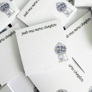 May include: White notepads with a gumball machine illustration and the text "just one more chapter" in black script. The gumball machine is gray with blue and purple gumballs. The notepads are stacked.