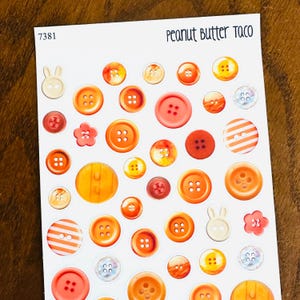 May include: A sheet of stickers featuring various orange and red buttons in different sizes and styles. Some buttons have four holes, others have two. There are also two bunny-shaped and two flower-shaped stickers. The text "Peanut Butter Taco" is at the top.