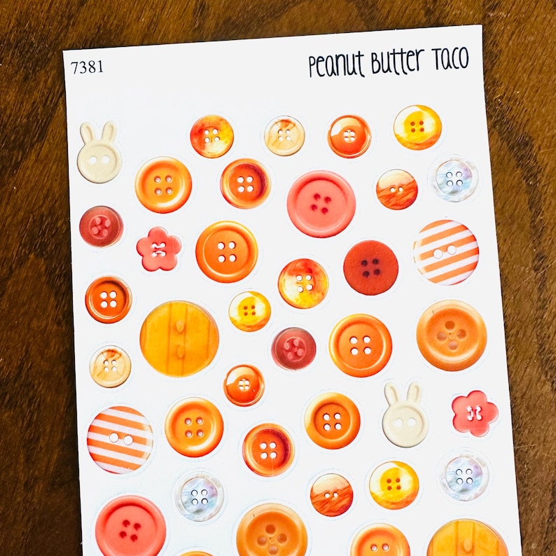 May include: A sheet of stickers featuring various orange and red buttons in different sizes and styles. Some buttons have four holes, others have two. There are also two bunny-shaped and two flower-shaped stickers. The text "Peanut Butter Taco" is at the top.