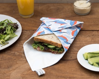 May include: A white reusable food wrap with a blue and orange bird pattern is folded around a sandwich. The sandwich is on a wooden table with a glass of orange juice, a bowl of hummus, and a plate of cucumber slices.
