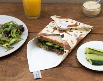 May include: A reusable sandwich wrap with a deer pattern, holding a sandwich with lettuce and cucumber. A glass of orange juice, a plate of salad, and a small bowl of hummus are also on the wooden table. The wrap is beige with brown deer.