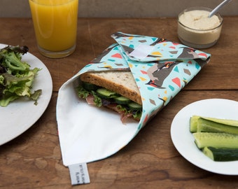 May include: A reusable sandwich wrap with a teal background and a penguin pattern. The wrap is open, revealing a sandwich with cucumber and lettuce. A glass of orange juice, salad, and hummus are also on the wooden table.