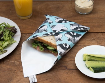 May include: A reusable sandwich wrap with a light blue background and a pattern of black and white badger faces. The wrap is open, revealing a sandwich with lettuce, cucumber, and ham. A glass of orange juice and a plate of salad are also visible.