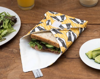 May include: A yellow and black patterned fabric wrap with a badger design, used to wrap a sandwich. The wrap is folded over a sandwich with lettuce, cucumber, and ham.