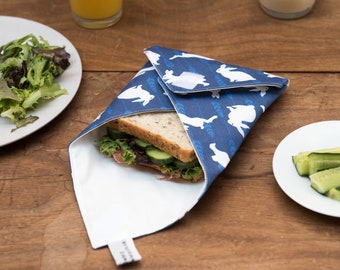 May include: A blue sandwich wrap with a white rabbit pattern, holding a sandwich with lettuce and cucumber. A glass of orange juice, a plate of salad, and a small bowl of hummus are on a wooden table.