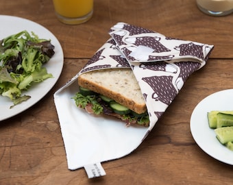 May include: A white fabric sandwich wrap with a brown bear print pattern. The wrap is folded around a sandwich with lettuce, cucumber, and ham. The wrap has a label that says "Reusable & Washable".