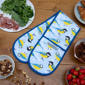 May include: A pair of blue oven mitts with a white background and a pattern of bluebirds with yellow accents. The mitts are made of fabric and have a blue trim. The mitts are on a wooden table with other items, including a bowl of nuts, a bowl of strawberries, and a bowl of granola.