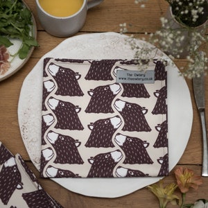 May include: A set of bear-themed napkins on a white plate, with a matching pattern. The napkins are beige with a repeating pattern of brown bear faces. The plate is set on a wooden table, with a fork and knife, and a small plate of food.