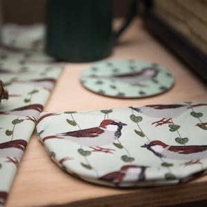 May include: A light green fabric set featuring a sparrow and leaf pattern. The set includes a coaster, a small pouch, and a napkin. The sparrow design has brown, white, and gray details. The items are on a wooden surface.