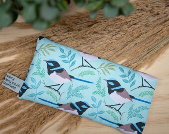 May include: A rectangular, light blue fabric item with a repeating pattern of stylized birds and foliage. The birds are black, blue, and white. The item has a small tag with text. The background includes dried grasses and greenery.