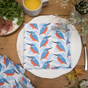 May include: White square cloth napkins with a repeating pattern of blue and orange kingfisher birds. The napkins are placed on a white plate, with a fork and knife, and other food items on a wooden table.