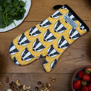 May include: A yellow oven glove featuring a repeating pattern of badger faces in black and white. The glove has a black trim and a hanging loop. The text "The Owerby" is visible. The glove is surrounded by food items on a wooden surface.