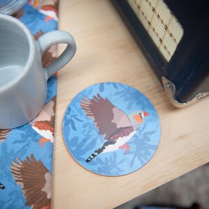 May include: A round coaster with a bird design. The coaster features a brown, white, and orange bird with outstretched wings against a light blue background with a leaf pattern. A blue mug and a patterned napkin are also visible.