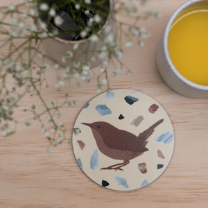 Peut inclure: Un dessous de verre circulaire illustré d'un troglodyte brun centré sur un fond crème. Le motif est agrémenté de formes géométriques multicolores éparpillées dans des tons de bleu, gris et brun, rappelant de petits fragments de pierre.