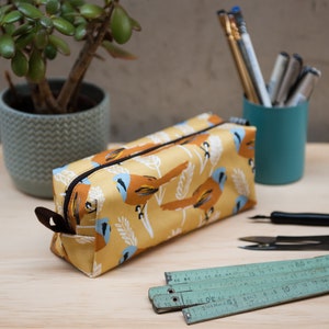 Könnte beinhalten: Ein rechteckiges Federmäppchen mit gelbem Hintergrund und einem Vogelmuster in Braun und Blau. Das Mäppchen hat einen schwarzen Reißverschluss und einen braunen Leder-Zuglasche. Weitere Artikel sind ein grüner Zollstock, Stifte und eine kleine Topfpflanze.