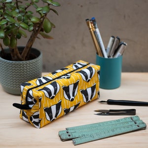 Könnte beinhalten: Gelbes Federmäppchen mit einem sich wiederholenden Muster aus schwarz-weißen Dachsgesichtern. Das Etui hat einen schwarzen Reißverschluss und einen Zipper. Weitere Gegenstände auf der Holzoberfläche sind eine kleine Topfpflanze, Stifte, Bleistifte und ein türkisfarbener Becher.