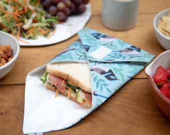 May include: A reusable sandwich wrap with a blue bird and leaf pattern, holding a sandwich with lettuce, tomato, and meat. The wrap is open, revealing the sandwich. Other food items, including a salad, grapes, and nuts, are in the background.