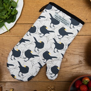 以下が含まれることがあります： 白地に黒い鳥の模様が入ったオーブンミトン。鳥は黄色いクチバシで、灰色の枝の背景に配置されています。ミトンは黒いトリムと吊り下げ用のループが付いています。「The Owlery」の文字が見えます。