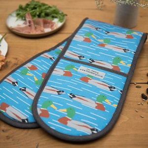 May include: Blue oven gloves featuring a duck design. The gloves have a light blue backdrop with white lines and duck illustrations. The edges are trimmed with grey fabric. The label reads "The Owlery www.theowlery.co.uk".