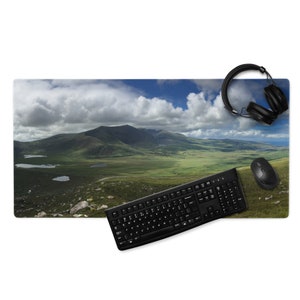 May include: A rectangular desk mat featuring a scenic landscape of green hills, lakes, and mountains under a cloudy sky. A black keyboard, mouse, and headphones are on the mat.
