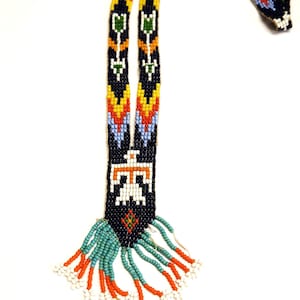 Puede incluir: Un collar largo de cuentas con un patrón geométrico en tonos de azul, naranja, amarillo y blanco. El collar tiene una franja de cuentas turquesa y naranja en la parte inferior.