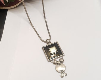 May include: A silver chain necklace with a square pendant. The pendant is black with a gold-colored center and has a silver charm hanging from it.