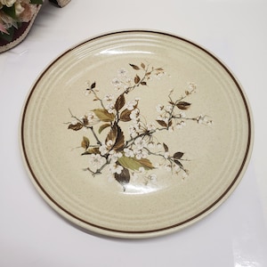 May include: A beige ceramic dinner plate with a brown rim and a floral design featuring white flowers and green leaves.