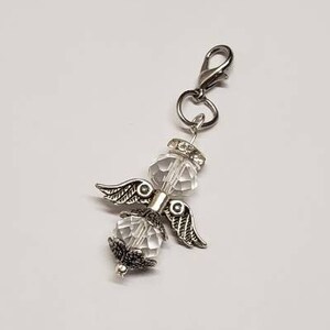May include: A silver charm featuring a clear crystal angel design. The charm has a silver clip at the top for attaching to a bracelet or necklace.