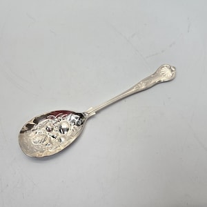 Może przedstawiać: Srebrna łyżka do serwowania z ozdobnym wzorem przedstawiającym grono winogron i liście. Łyżka ma zaokrągloną miseczkę i długi, smukły trzonek.