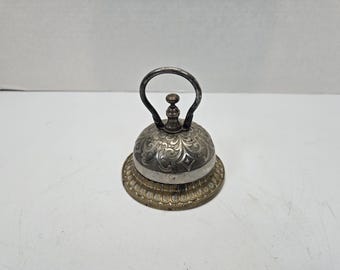 May include: A silver and gold decorative bell with an ornate design and a handle. The bell has a round base and a domed top.