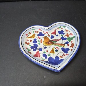 Puede incluir: Plato de cerámica en forma de corazón con un diseño pintado a mano. El plato presenta un interior blanco con un borde azul. El diseño incluye un pájaro colorido, flores azules y otros elementos florales. Una pieza decorativa.