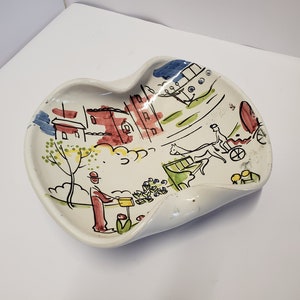 Puede incluir: Un plato de cerámica blanca con una escena pintada de fantasía de una ciudad con edificios, árboles y un carruaje tirado por un caballo. El plato tiene una forma ondulada y orgánica.